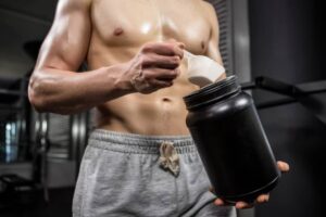 A fit man in gym shorts scoops protein powder from a black container, preparing for his workout session.