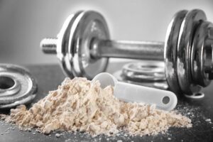 A white scoop rests beside a pile of protein powder, with dumbbells blurred in the background, symbolizing fitness and nutrition.