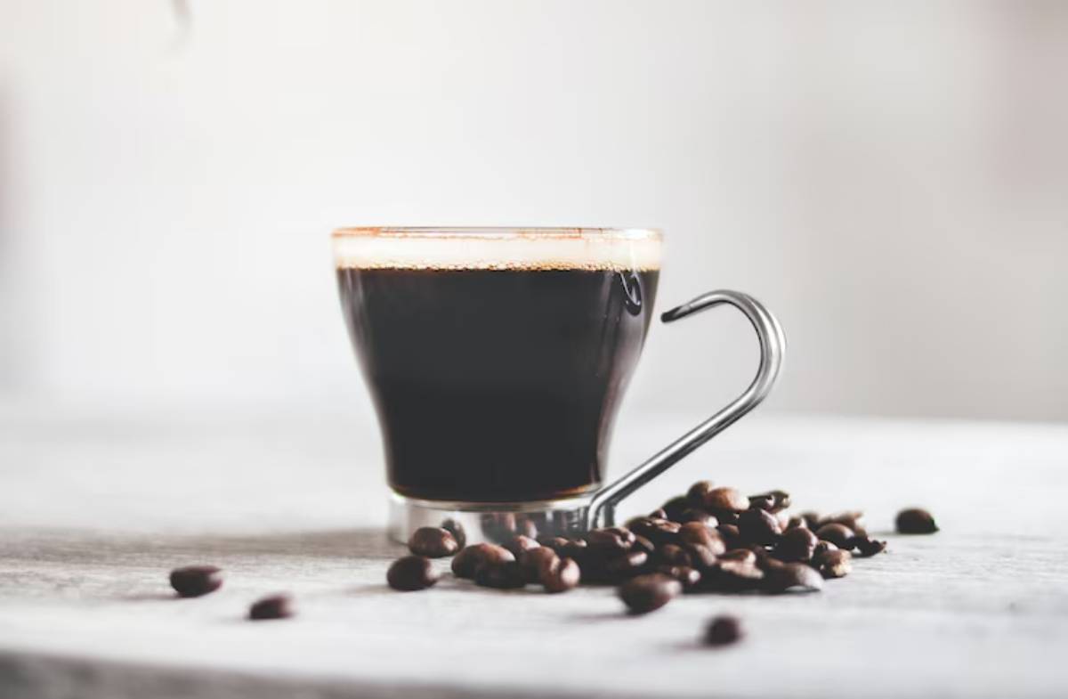 A stylish glass cup filled with dark coffee sits on a light surface, accompanied by scattered coffee beans.