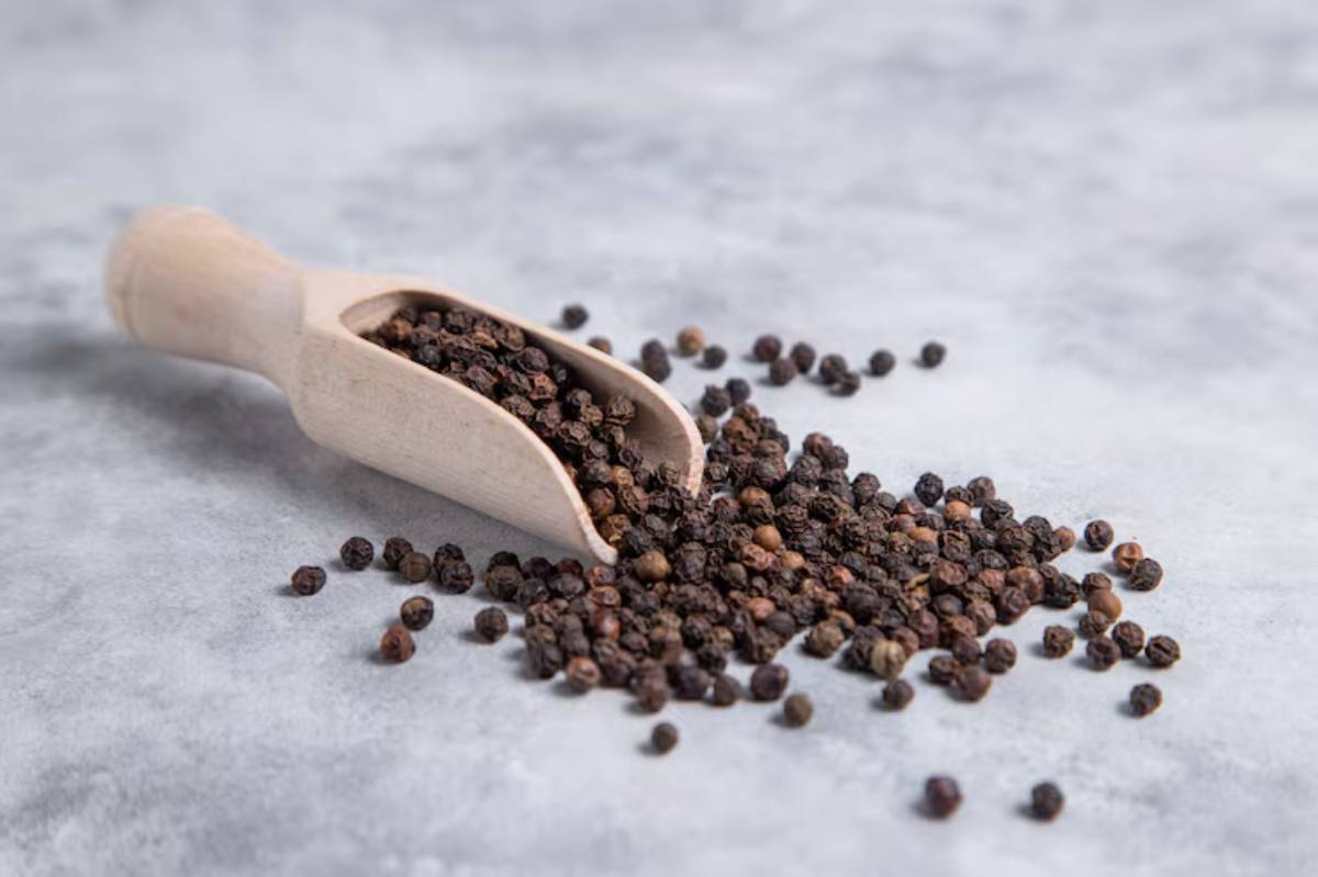 A wooden scoop filled with black peppercorns, some spilling onto a gray surface, highlighting their rich color and texture.