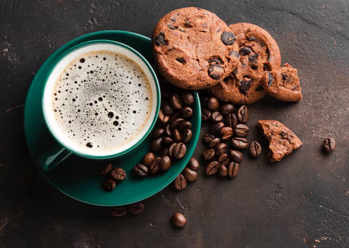 A teal cup of frothy coffee sits beside chocolate chip cookies and scattered coffee beans on a dark surface.