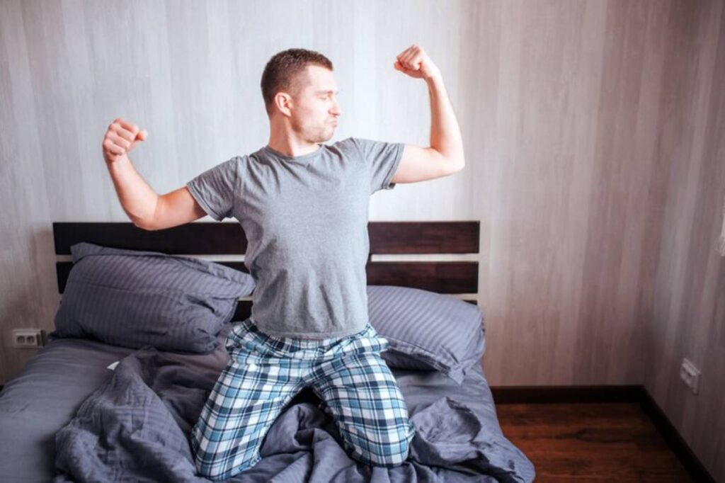 A person in pajamas looks triumphant while kneeling on a bed, surrounded by cozy gray bedding in a softly lit room.