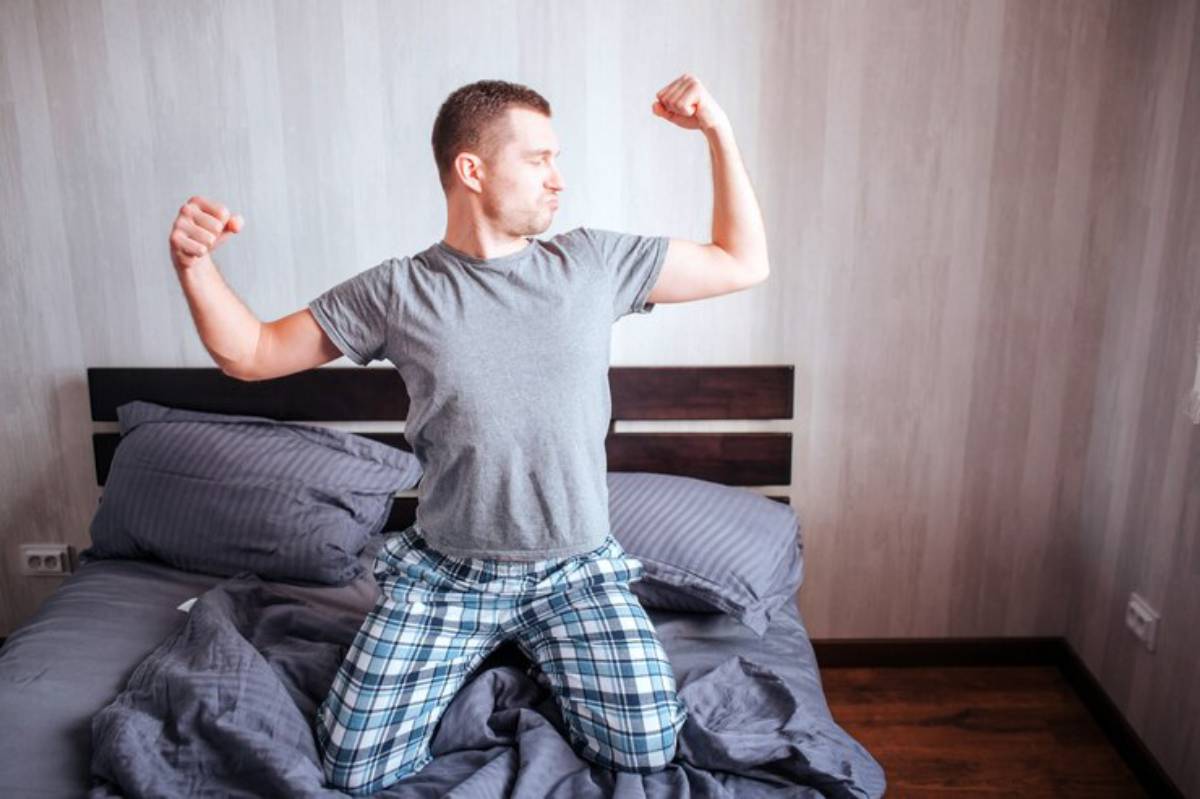 A person in pajamas looks triumphant while kneeling on a bed, surrounded by cozy gray bedding in a softly lit room.