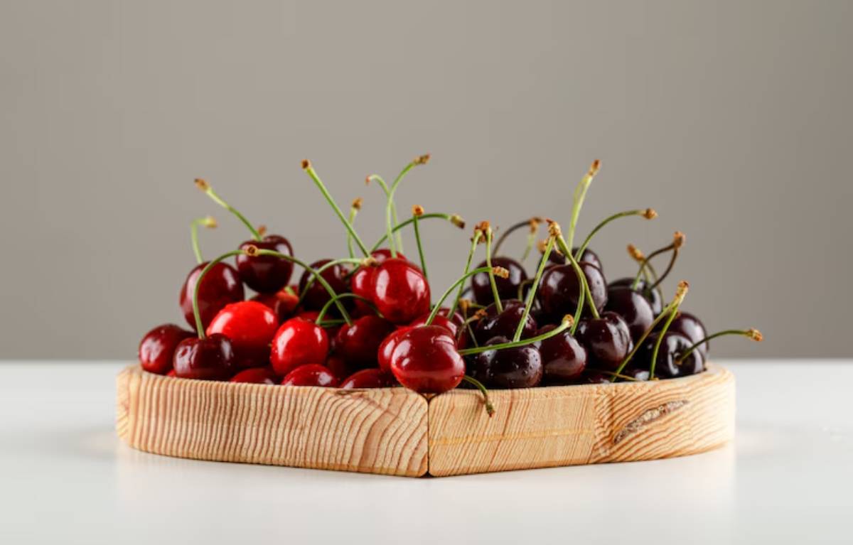 A wooden bowl filled with fresh red and dark purple cherries, resting on a light surface against a simple gray background.