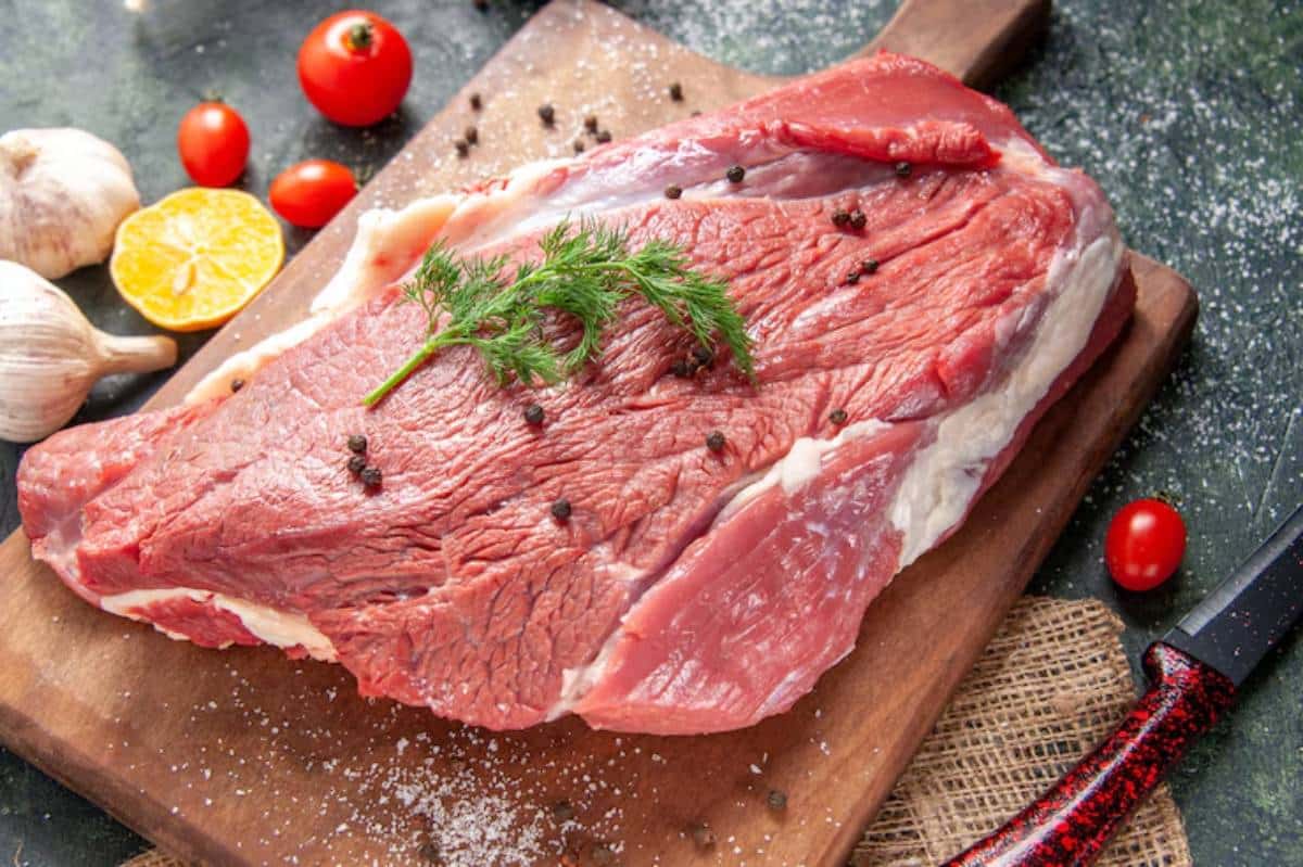 Raw red meat with a sprig of dill, surrounded by garlic, cherry tomatoes, and a slice of lemon on a wooden cutting board.