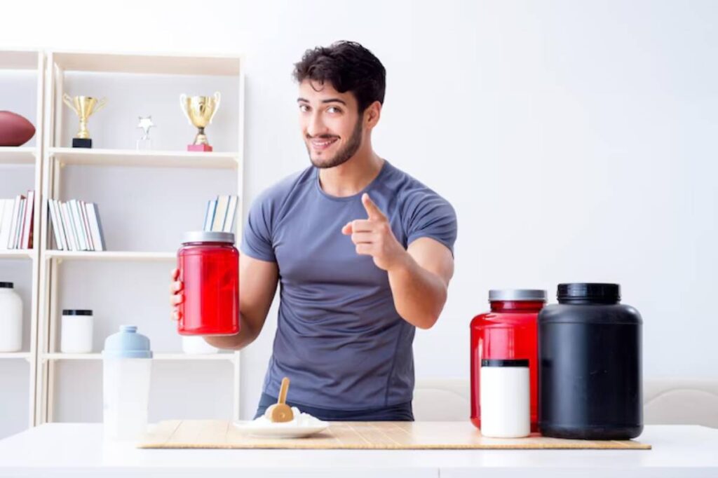 A person in a gray shirt stands behind a table with various supplement containers, pointing while holding a large red jar.