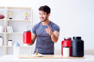 A person in a gray shirt stands behind a table with various supplement containers, pointing while holding a large red jar.