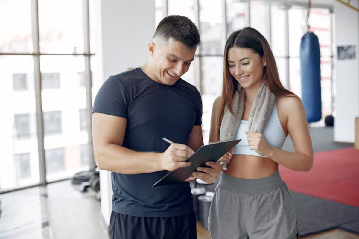 A trainer assists a female client with fitness guidance in a sunny gym, surrounded by equipment and windows.