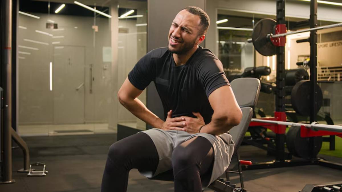 A person in athletic clothing sits on a gym bench, holding their stomach with a pained expression, surrounded by gym equipment.