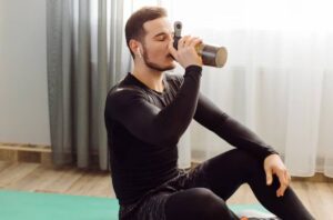 A fit individual in athletic wear sits on a yoga mat, sipping from a shaker bottle, with sunlight streaming through sheer curtains.