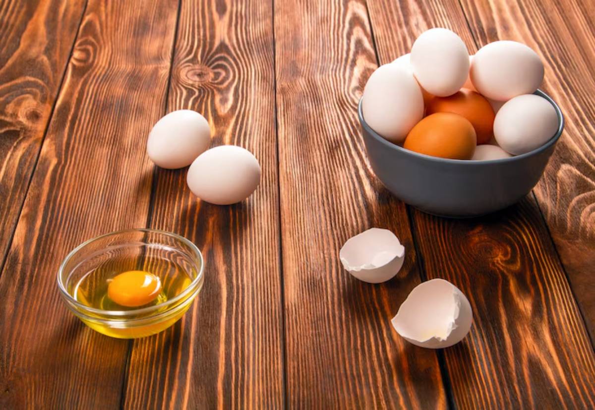 A bowl of white eggs with one brown egg, alongside a cracked shell and a bowl of raw egg, on a wooden table.