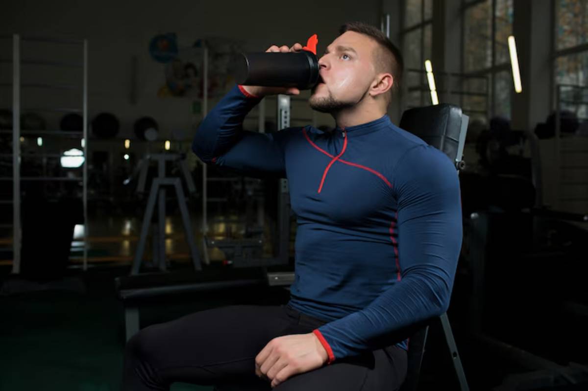 A muscular person in a blue athletic top sits on a gym bench, sipping from a black shaker bottle in a fitness environment.