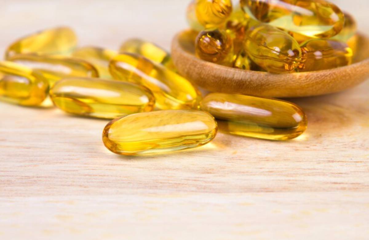 A close-up of golden gelatin capsules scattered on a wooden surface, with a small wooden bowl filled with more capsules.