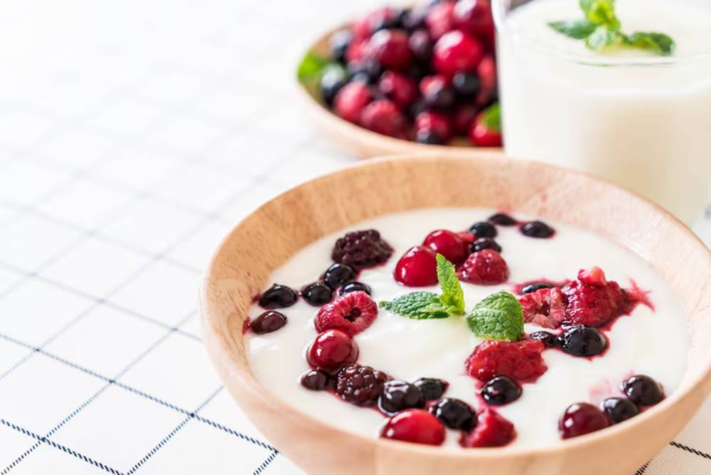A wooden bowl filled with creamy yogurt topped with a variety of fresh berries and mint leaves, with another berry bowl in the background.