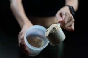A close-up of a hand holding a scoop of protein powder above a container against a dark background.
