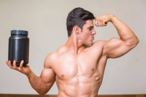 A muscular man flexes his bicep while holding a large black container of protein powder, showcasing his fitness dedication.