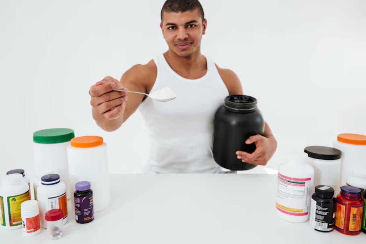 A person in a white tank top holds a spoonful of powder with a black container while surrounded by various supplement bottles on a table.