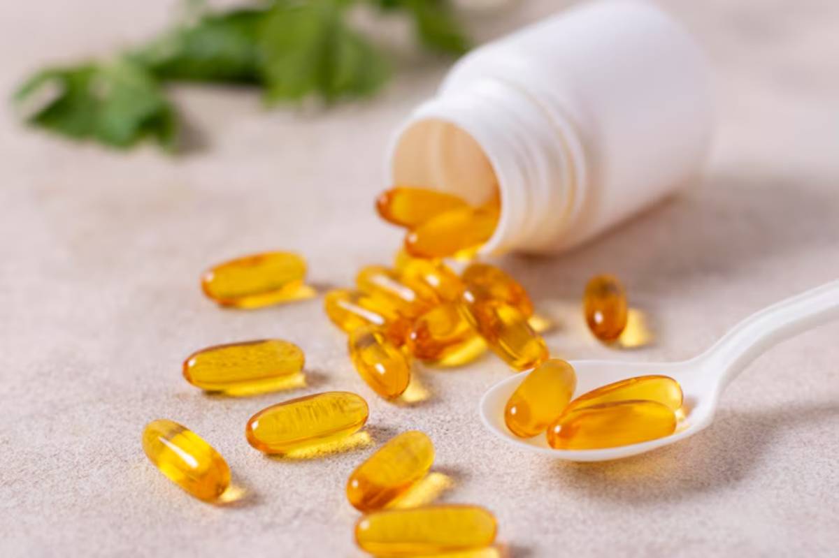 A white bottle spills golden gel capsules onto a light surface, with a small spoon holding some of the capsules nearby.