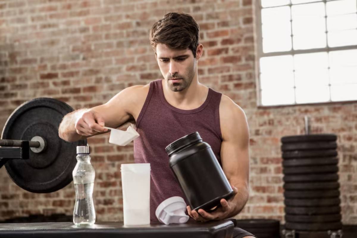 A muscular individual in a tank top measures protein powder into a shaker bottle, surrounded by gym equipment and a water bottle.