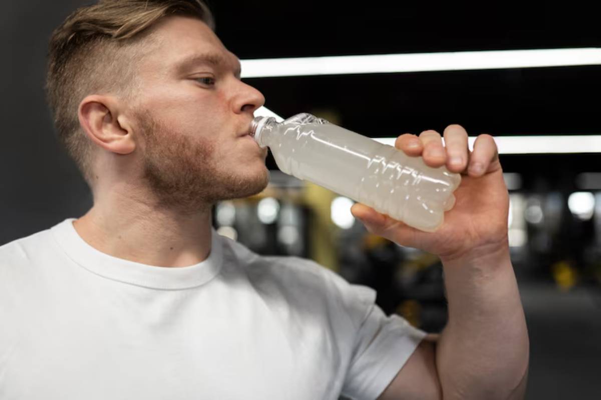 A person in a white t-shirt drinks from a plastic bottle filled with a cloudy liquid, set in a gym environment.