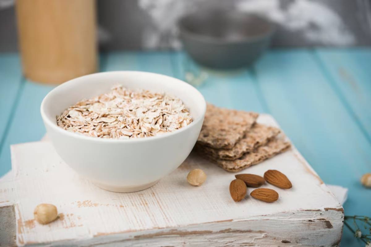 A white bowl filled with oats sits atop a rustic wooden board, accompanied by nuts and whole-grain crackers on a blue background.