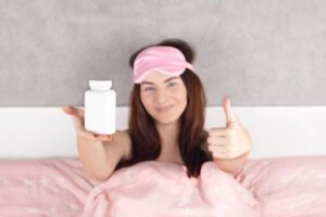 A person in bed holding a bottle of pills with a thumbs-up sign, wearing a pink sleep mask. Cozy bedroom setting.