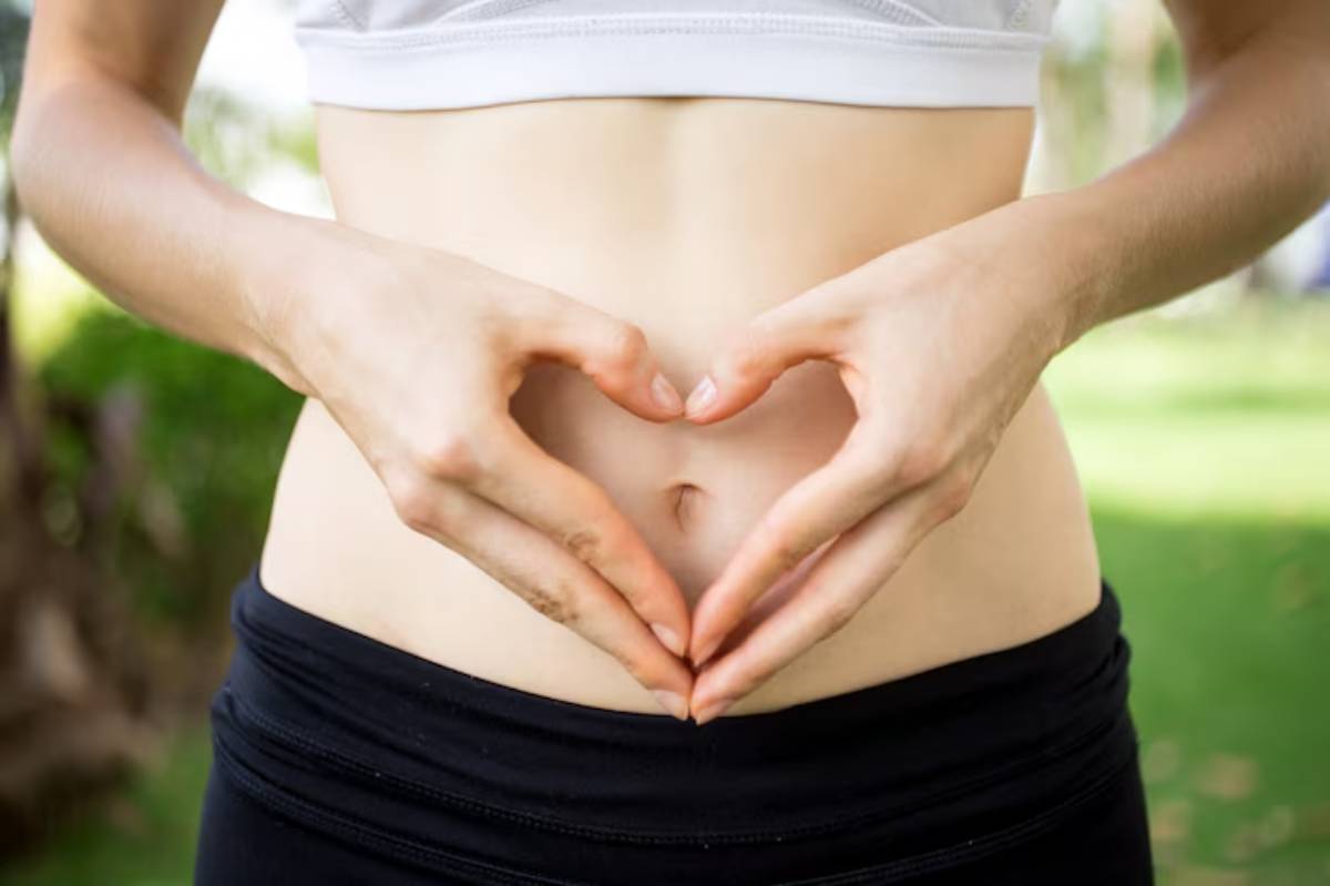A person in athletic wear forms a heart shape with their hands over their abdomen, set against a green, outdoor background.