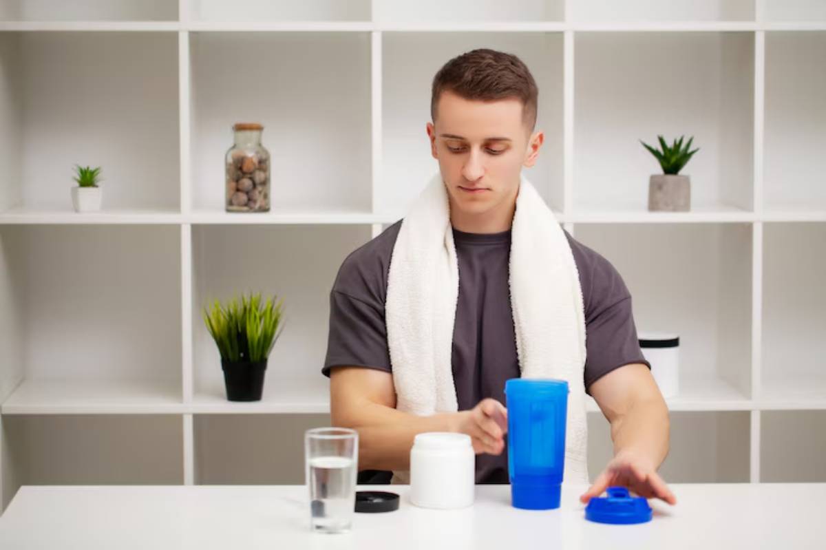 A person sits at a table with a blue shaker bottle, white protein powder container, and a glass of water, surrounded by simple decor.