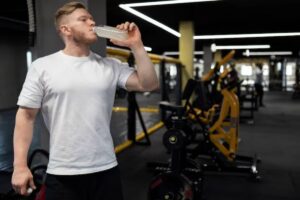 A person in a white t-shirt drinks from a clear bottle in a modern gym featuring exercise equipment in the background.