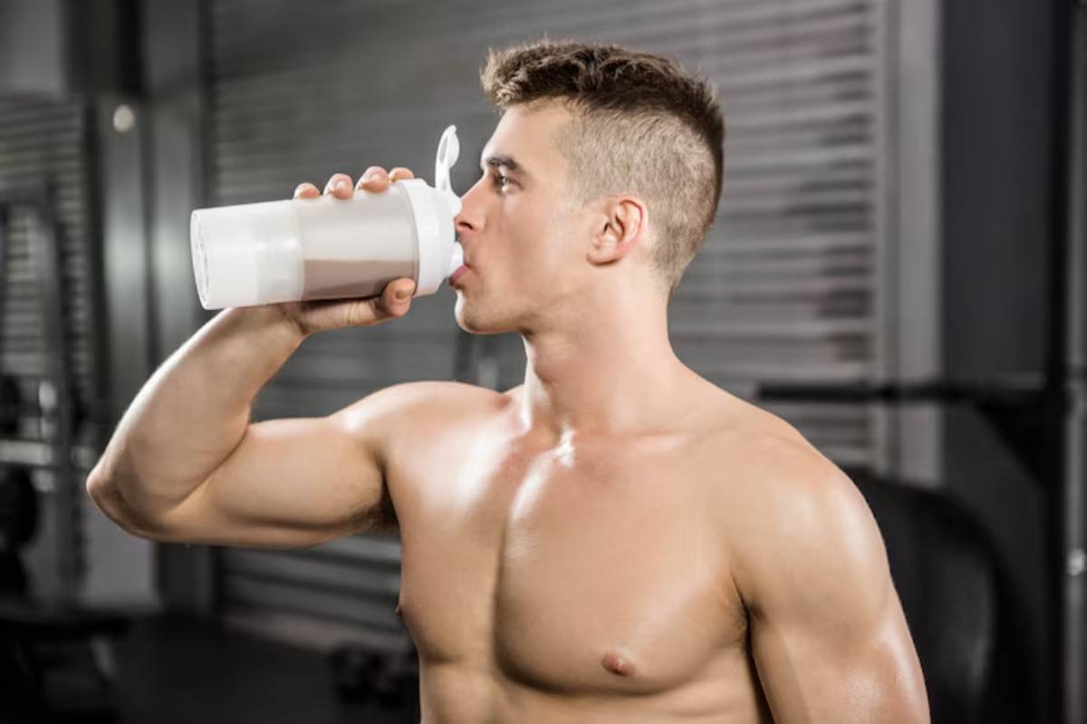 A shirtless man is drinking from a shaker cup in a gym setting, showcasing his muscular arm against blurred gym equipment.