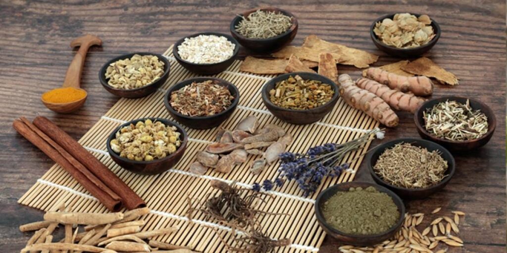 A variety of spices and herbs displayed in small bowls on a bamboo mat, including turmeric, cinnamon, and dried flowers.