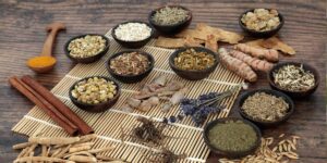 A variety of spices and herbs displayed in small bowls on a bamboo mat, including turmeric, cinnamon, and dried flowers.
