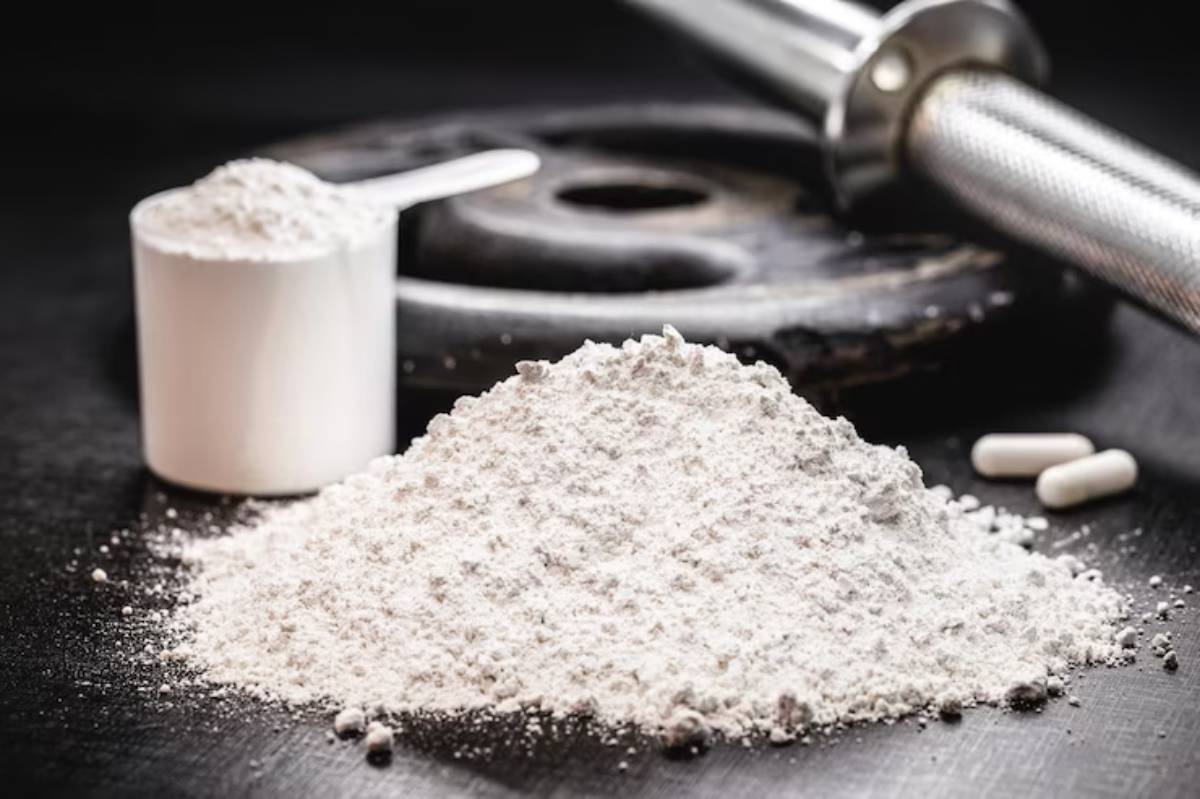A heap of white powder with a measuring scoop nearby, alongside an exercise weight plate and two capsules on a dark surface.