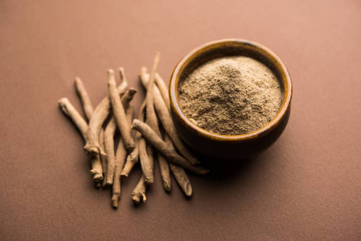A small wooden bowl filled with fine, light brown ashwagandha powder, beside several dried ashwagandha roots on a brown background.