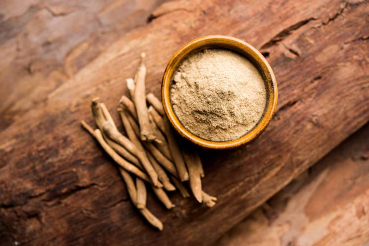 A wooden bowl of ashwagandha powder surrounded by dried ashwagandha roots, placed on a rustic wooden surface.