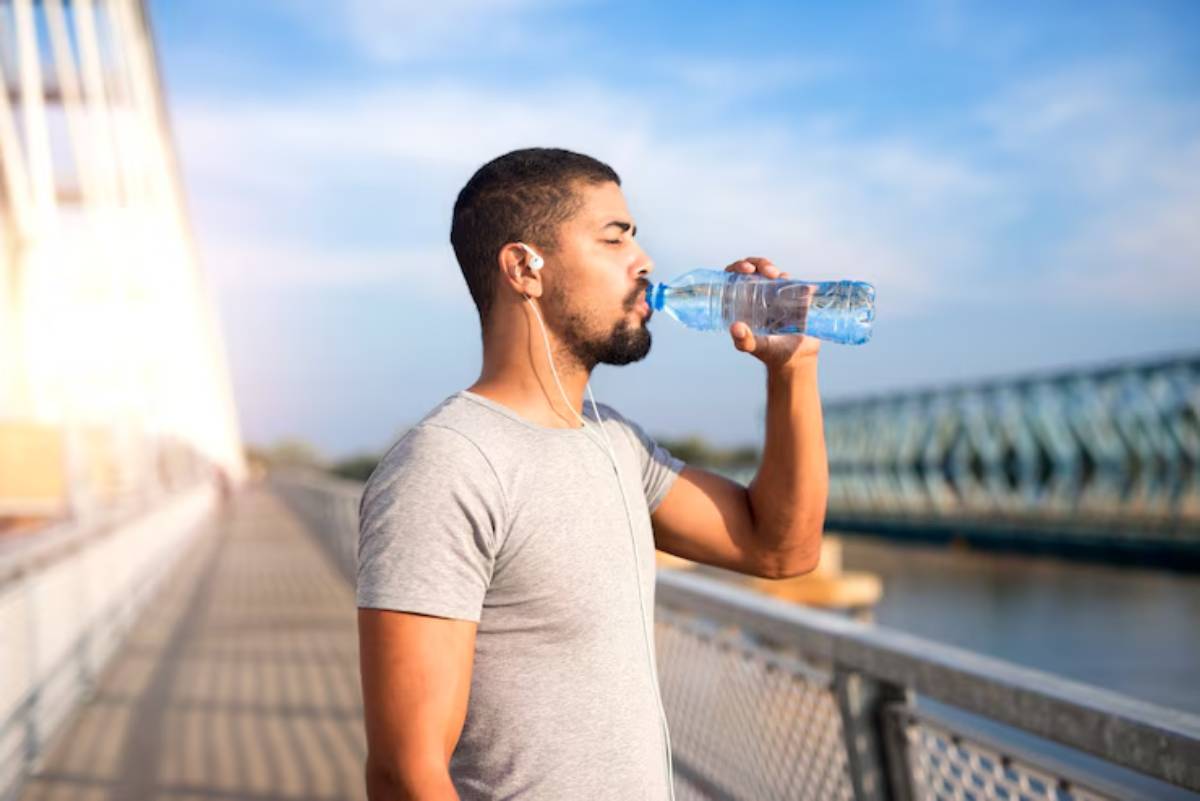 A man in a gray t-shirt drinks from a water bottle while standing on a bridge, with a scenic view of blue skies and water.