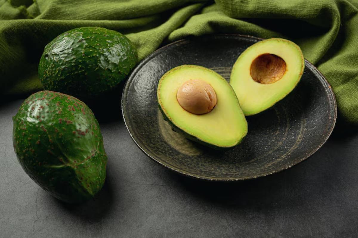 A plate with two halved avocados, one with its pit, beside whole avocados and a green cloth on a dark surface.