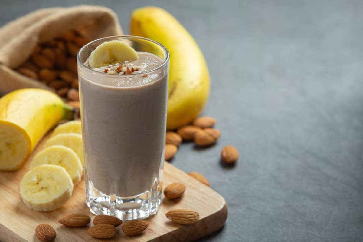 A glass of banana almond smoothie topped with a banana slice, surrounded by fresh bananas and scattered almonds on a wooden board.