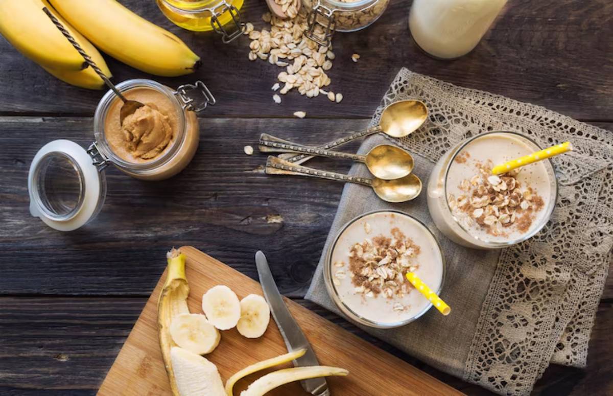 Two glasses of banana smoothie topped with oats, a jar of peanut butter, and fresh bananas on a rustic wooden table.
