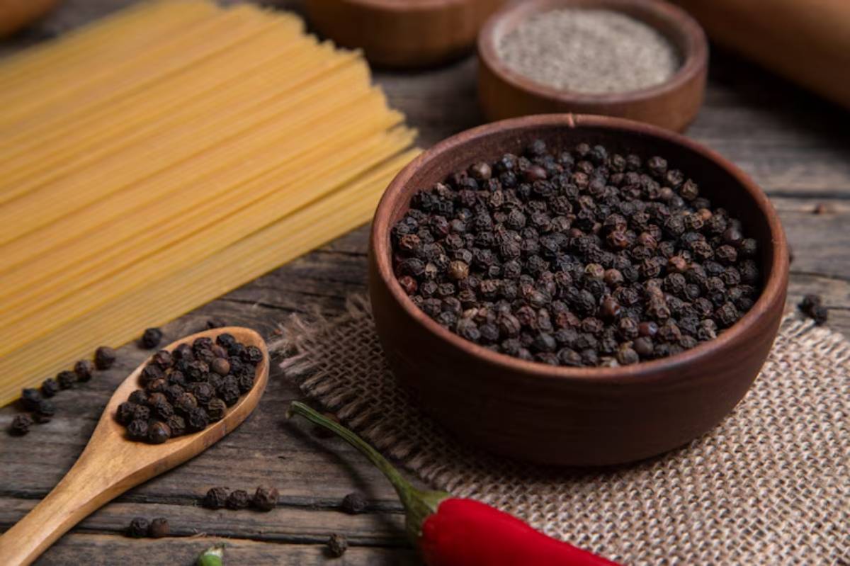 A wooden bowl filled with black peppercorns, alongside a spoonful of pepper, pasta, and a red chili pepper on a rustic table.