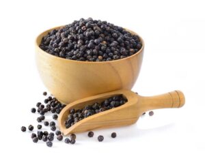 A wooden bowl filled with black peppercorns, accompanied by a wooden scoop and scattered peppercorns on a white background.