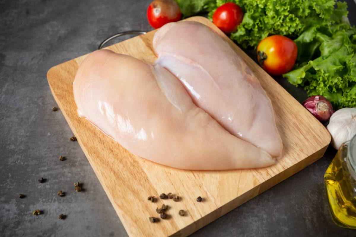 Raw chicken breasts on a wooden cutting board, surrounded by fresh vegetables, including lettuce, tomatoes, and garlic.