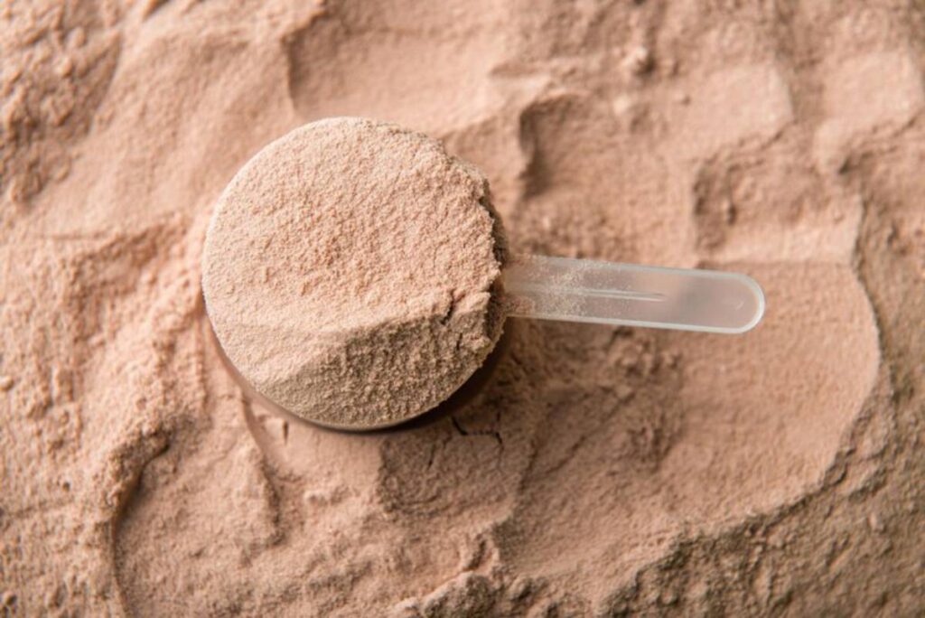 A close-up of a plastic scoop filled with a light brown powder, surrounded by loose powder on a textured surface.