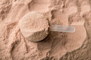 A close-up of a plastic scoop filled with a light brown powder, surrounded by loose powder on a textured surface.