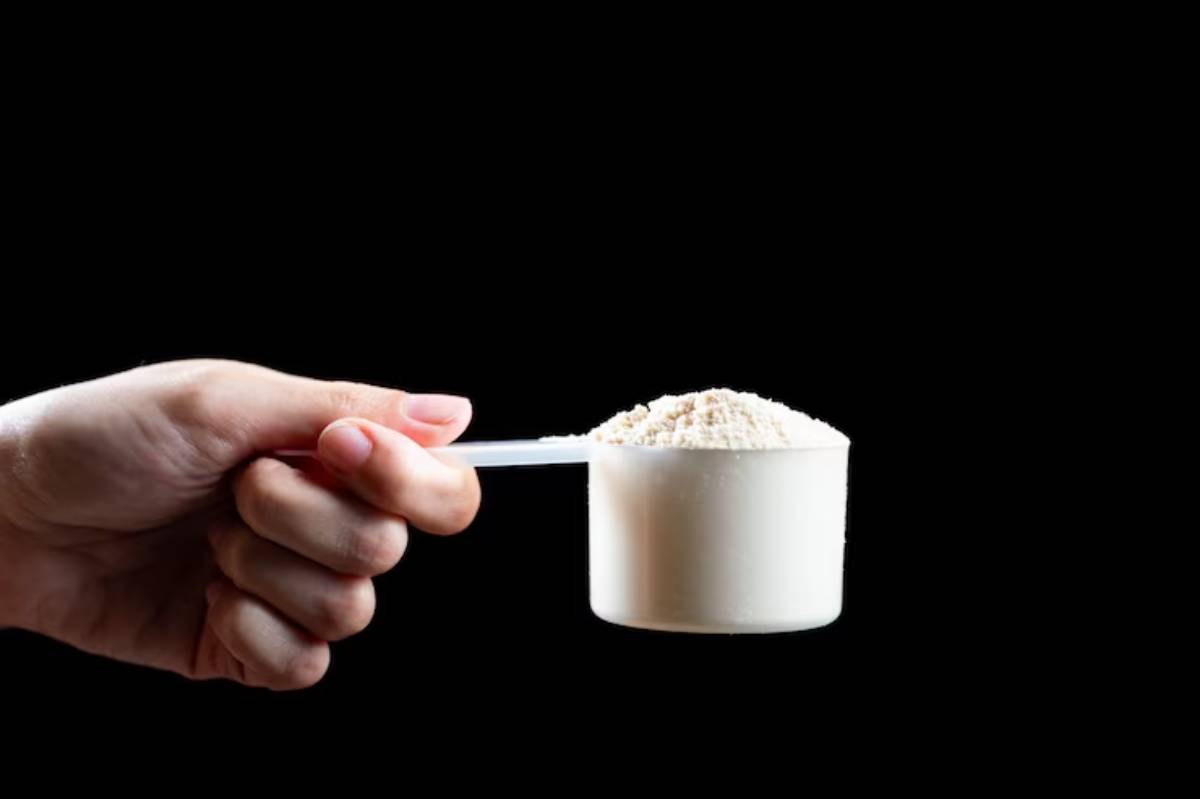 A hand holds a white plastic measuring scoop filled with a fine white powder against a black background.