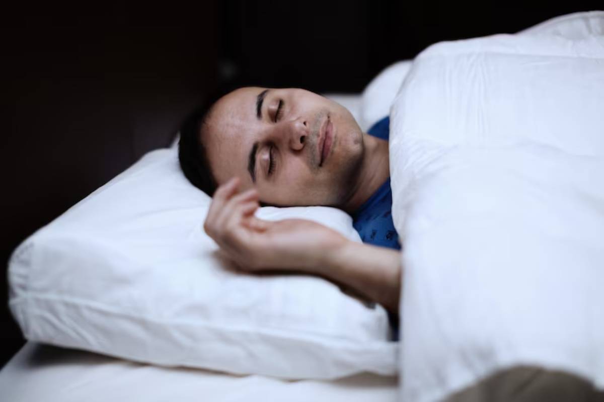 A person lies in bed covered with white sheets, resting their hand on the pillow, suggesting a peaceful sleep environment.