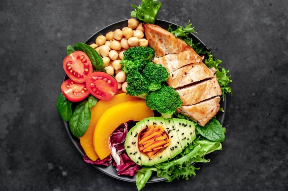 A vibrant salad plate featuring grilled chicken, broccoli, chickpeas, tomatoes, spinach, avocado, and colorful squash on a dark background.