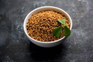 A white bowl filled with golden-brown fenugreek seeds, garnished with fresh green leaves, on a dark textured surface.
