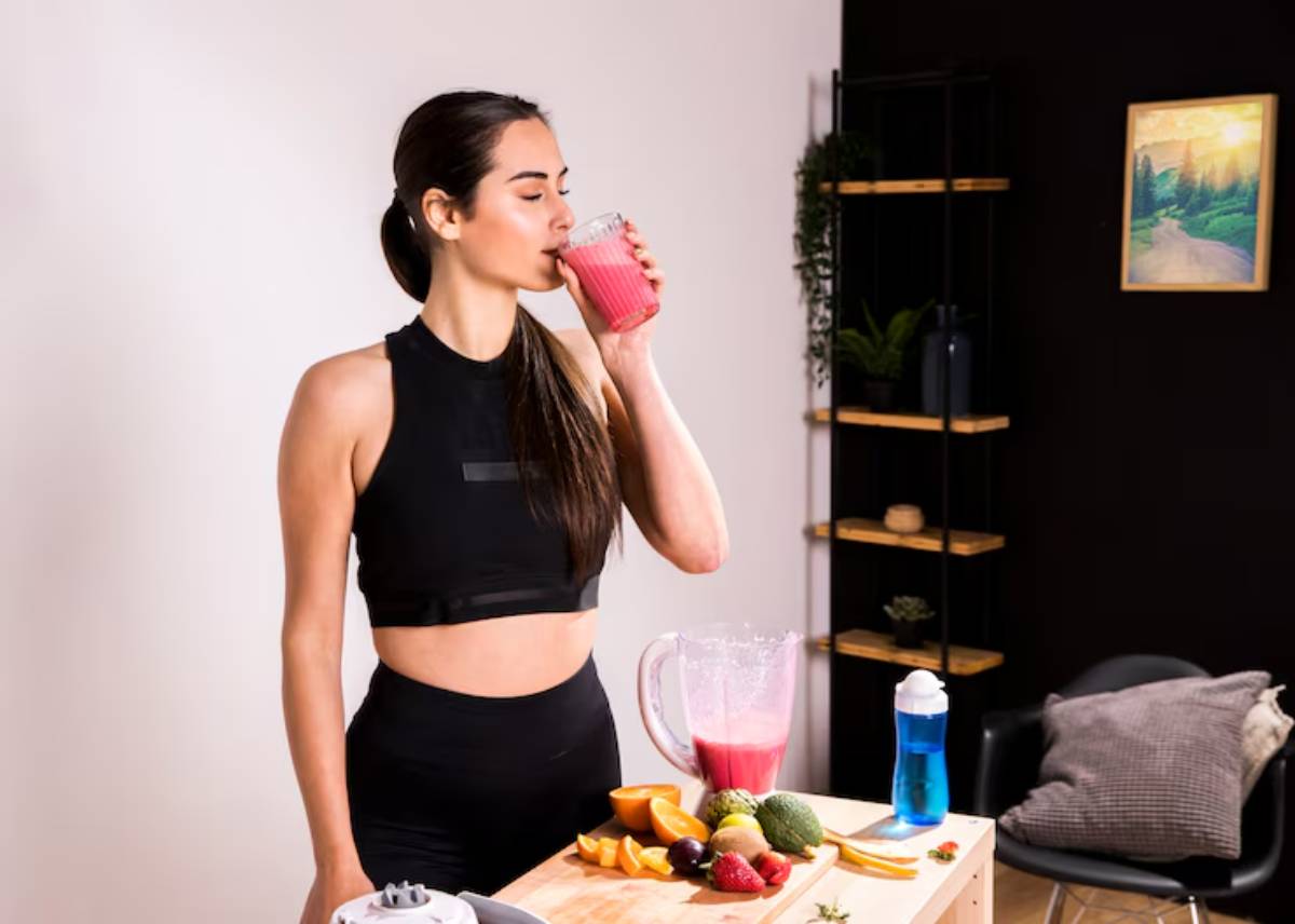 A woman in athletic wear drinks a pink smoothie, surrounded by fresh fruits and a blender in a modern kitchen setting.