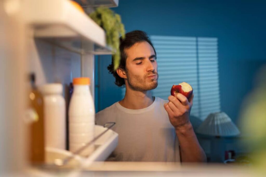 A person stands in front of an open refrigerator, holding a partially bitten red apple, surrounded by various foods and drinks.